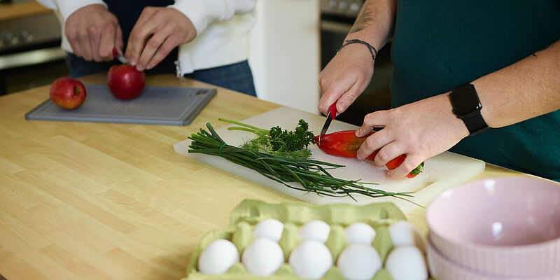 Zwei Personen und deren Hände sind zu sehen, wie sie verschiedenes Gemüse auf Brettern schneiden. Im Vordergrund ist ein offener Karton mit 10 Eiern zu sehen.