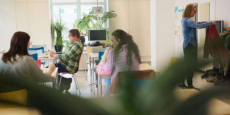 Auf dem Bild sind mehrere Menschen zu sehen welche an verschiedenen Tischen sitzen. Zwei Frauen unterhalten sich, ein Mann blickt auf sein Handy. Auf der rechten Seite des Bildes, hält eine Frau einen roten Pullover.